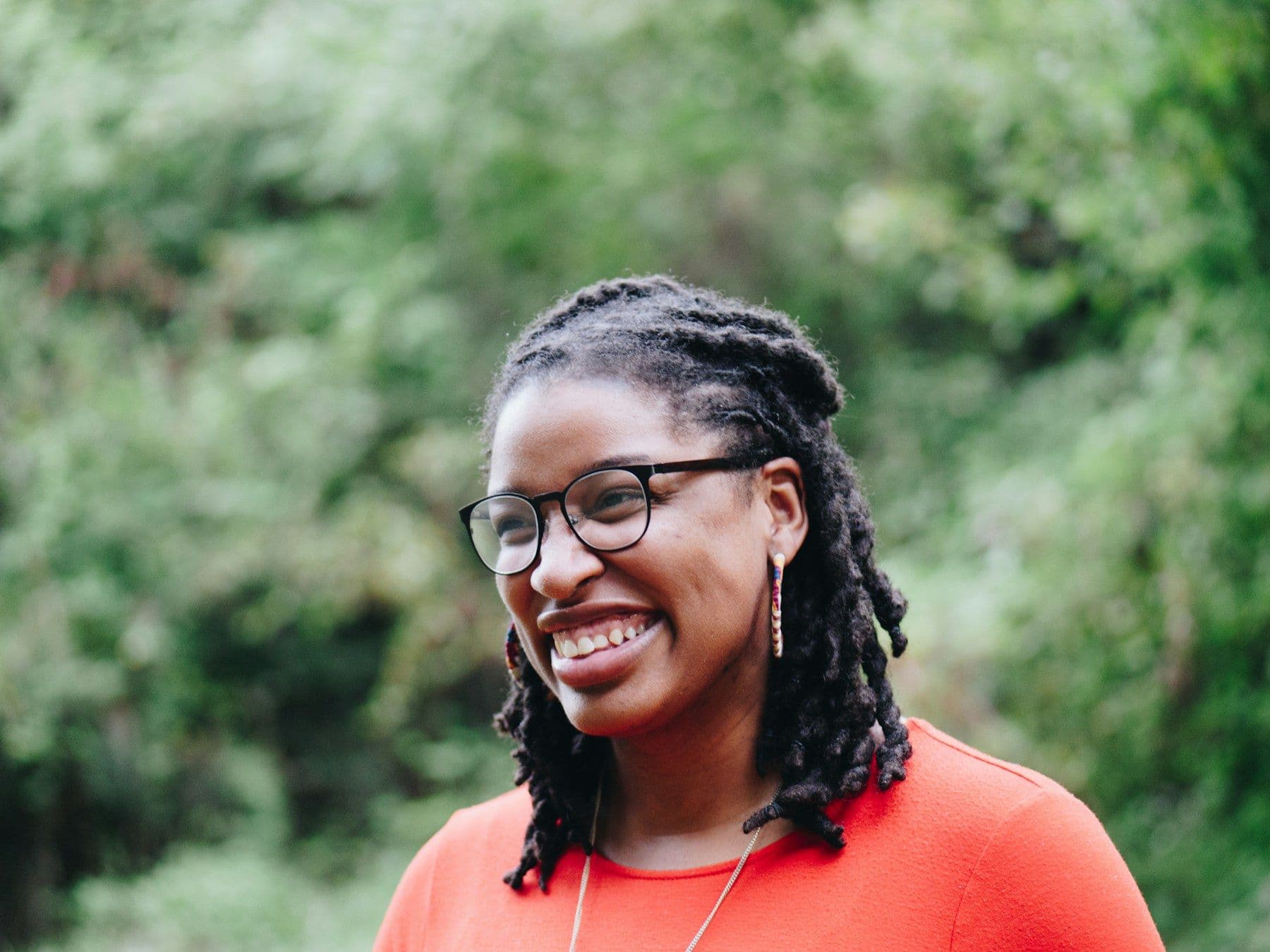 A smiling Black woman with glasses standing outdoors among trees